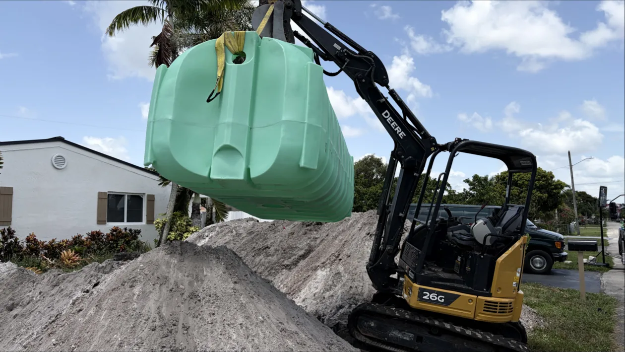 A green plastic septic tank hanging from a mini excavator over piles of sand beside a house, ready to be set in the ground
